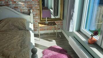 A bedroom with a brick wall, a bed with a patterned pillow, a small table with a lamp, a window with a view, and a radiator.