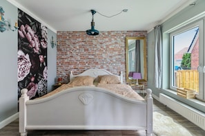 Ein Schlafzimmer mit einem weißen Bett, einer Ziegelwand, einem großen Fenster und einer Wandmalerei mit floralen Motiven.