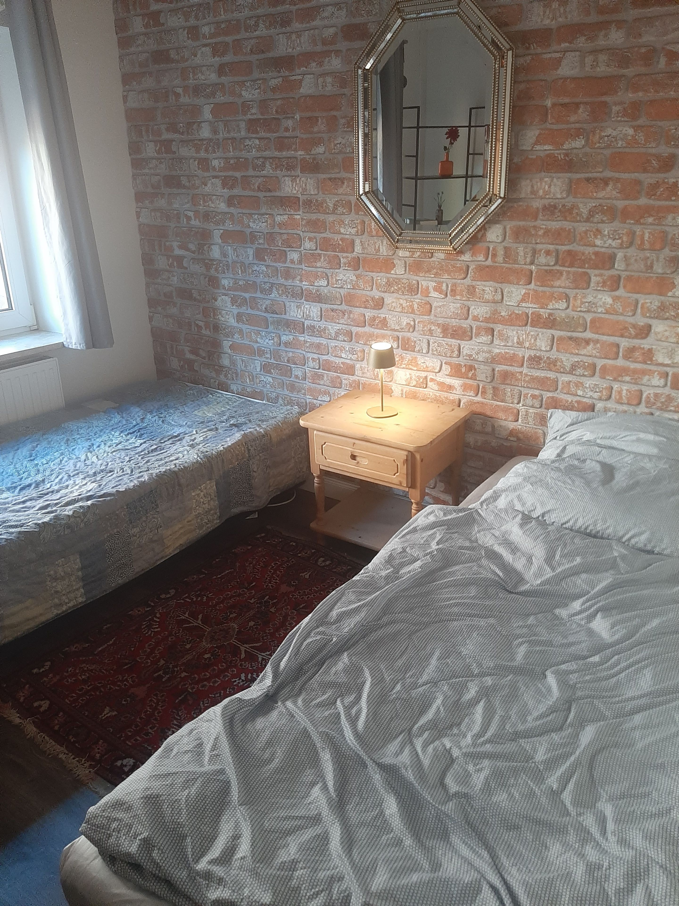 Quarto com parede de tijolos, cama com colcha branca, mesa de cabeceira com abajur e janela com cortinas.
