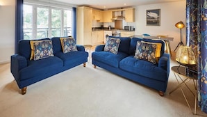 Een woonkamer met twee blauwe banken, een kleine salontafel en een eettafel met stoelen. Achtergrond toont een keuken met houten kastjes en een magnetron.