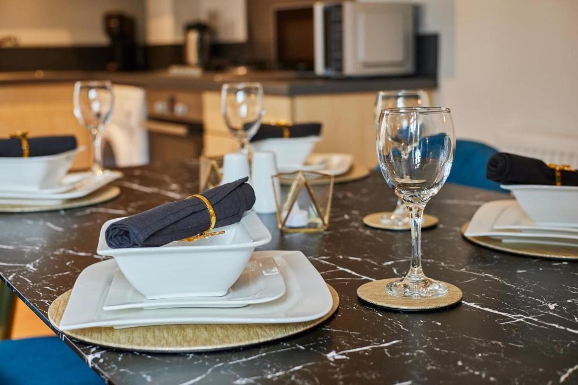 A dining table set with white plates, folded napkins, and clear wine glasses, with a modern kitchen in the background.