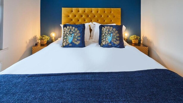 Een bed met een gestoffeerd hoofdeinde, twee decoratieve kussens met pauwendesigns, en een donkerblauwe plaid.