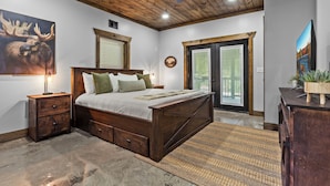Un dormitorio con techo de madera, cama con cajones para guardar objetos, mesita de noche, un cuadro grande y vista a través de una puerta de cristal.