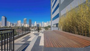 A rooftop terrace with a view of a city skyline, a wooden deck, and a bamboo plant. - Modern Studios in a Building With a Pool in Vila Cordeiro - CTB (São Paulo)
