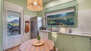 A kitchen with a round table, a hanging light fixture, a refrigerator, and a window with a view of trees. - Holiday Home 'Tordera Village' with Mountain View, Private Pool and Wi-Fi (Tordera)