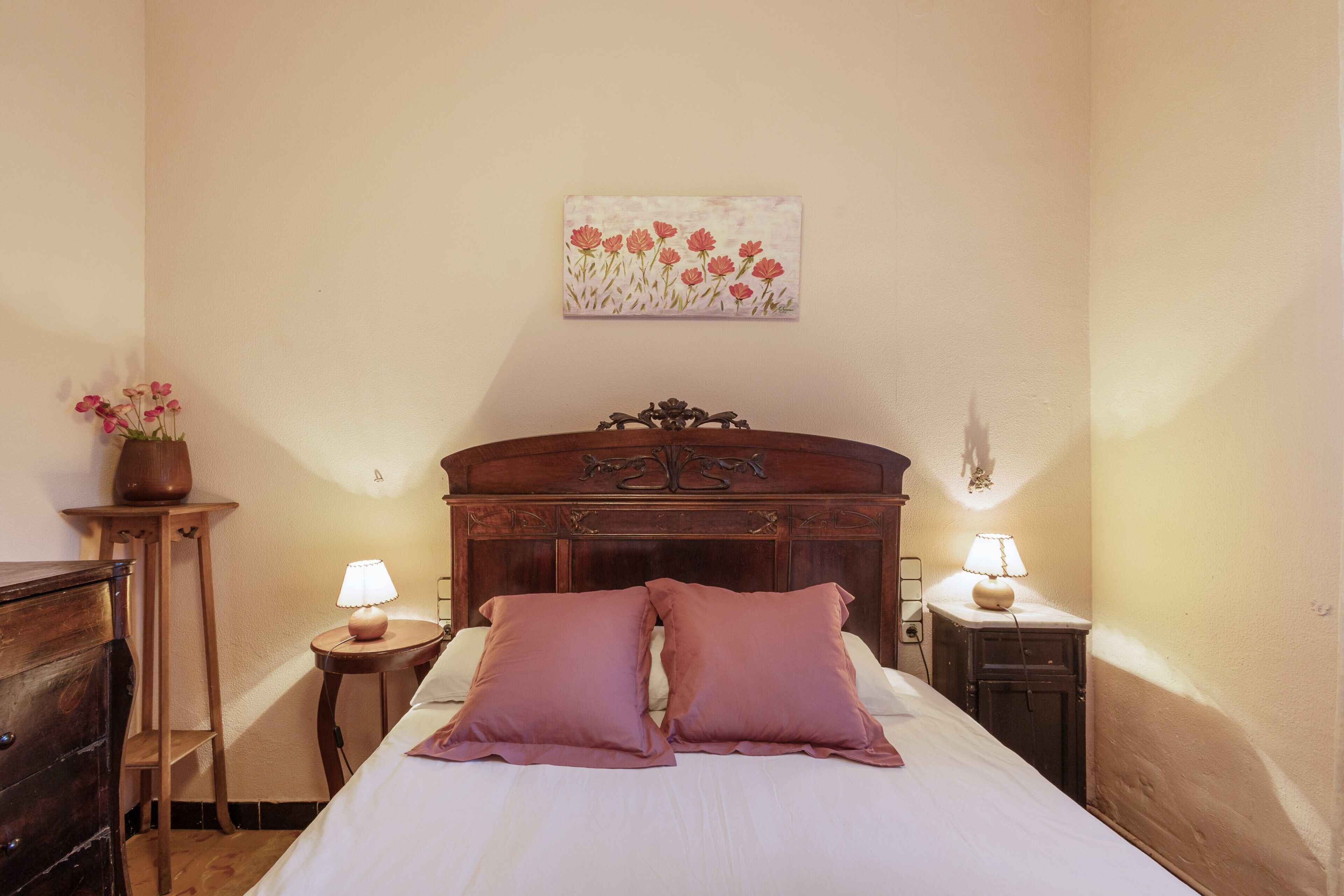 A bedroom with a wooden headboard, two bedside lamps, a small table with a vase, and a painting on the wall.