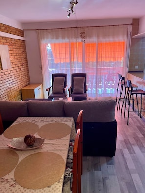 Ein Wohnzimmer mit einer Sofagarnitur, einem Esstisch mit Platzsets und Blick durch ein Fenster mit orangefarbenen Vorhängen.