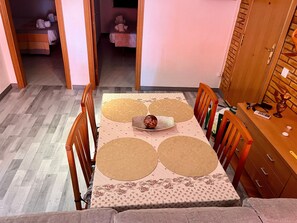 Un comedor con una mesa dispuesta para cuatro personas, rodeada de sillas de madera. En el fondo, una mesita con un objeto decorativo.