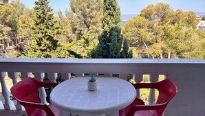 Un balcon avec une table blanche et deux chaises rouges, un pot de fleurs et une vue sur des arbres et une étendue d’eau.