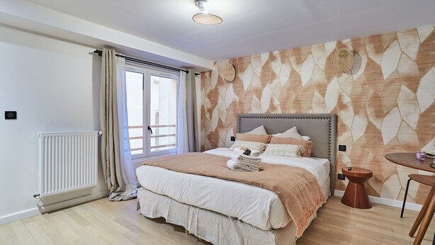 A bedroom with a bed, a bedside table, a window with curtains, a radiator, and a patterned wall.