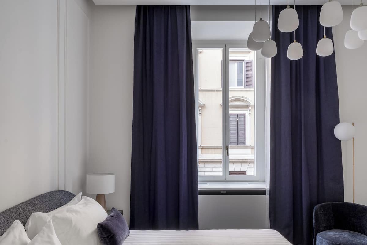 Ein modernes Hotelzimmer mit einem großen Bett, dunklen Vorhängen, Blick auf ein Gebäude durch das Fenster und einem Sessel.