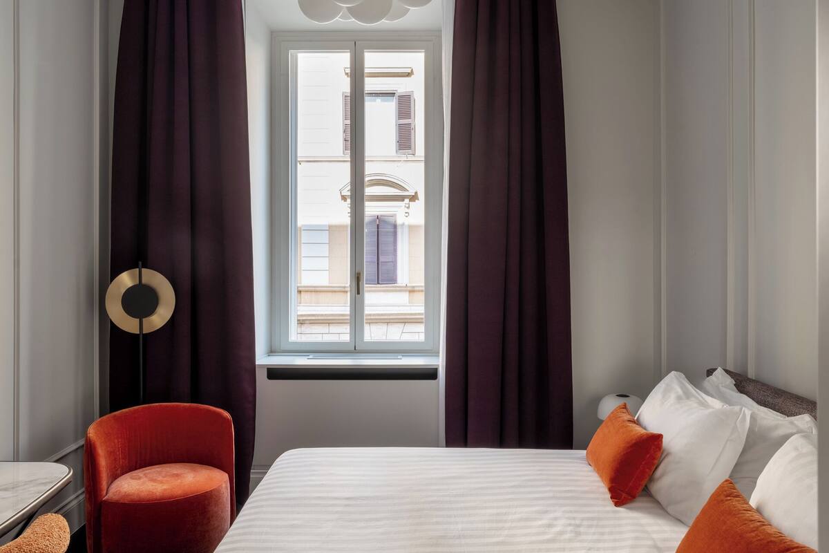 Ein Hotelzimmer mit Bett, Sessel, Stehlampe und einem Fenster mit Vorhängen.
