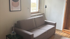 A modern living room with a grey sofa, a small side table with a bottle and glasses, a framed abstract artwork, a floor lamp, and a window with a view of greenery outside. - MYA Weinland Hotel Kobern-Gondorf (Kobern-Gondorf)