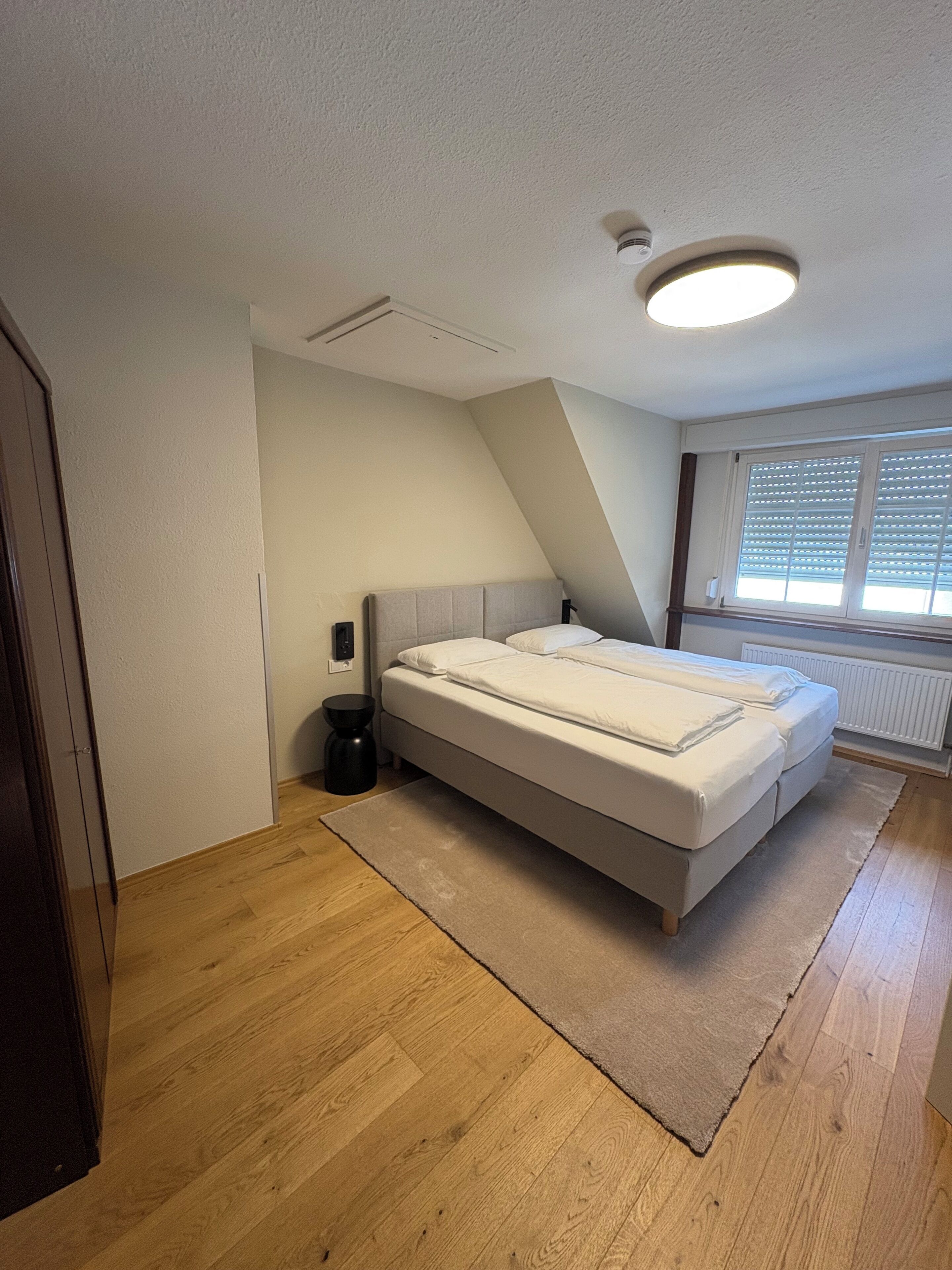 A bedroom with a bed, a radiator, a window with blinds, and a ceiling light.
