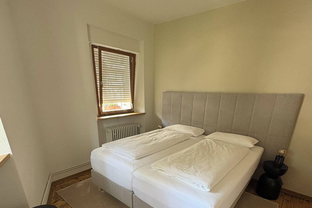 Ein Schlafzimmer mit Doppelbett, Fenster mit Jalousien, Heizkörper, Holzboden und einem Stuhl.