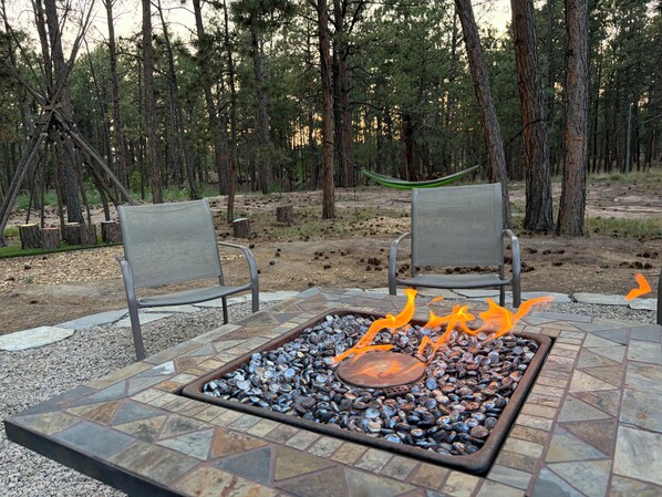 An outdoor seating area with a fire pit surrounded by stones, two metal chairs, and a forested backdrop.