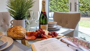 Ein Tisch mit einer Moët Champagnerflasche, Brot, Trauben und Wurstwaren, dazu ein Topfpflanze und ein Buch in einem Raum mit Blick auf Grünflächen.