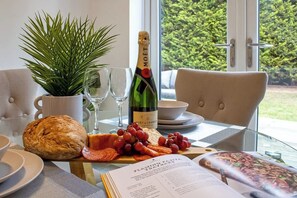 A table with a bottle of Moët champagne, a loaf of bread, grapes, and charcuterie, with a potted plant and a book on a table in a room with a view of greenery. - Beautiful & Cosy Home | Driveway | Close to M1 (Milton Keynes)