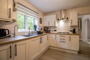 A kitchen with white cabinets, a gas stove, a coffee maker, and a window with blinds. - Beautiful & Cosy Home | Driveway | Close to M1 (Milton Keynes)