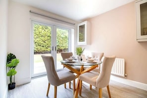 A dining area with a glass table, four chairs, a potted plant, and a view through a sliding door to a garden. - Beautiful & Cosy Home | Driveway | Close to M1 (Milton Keynes)