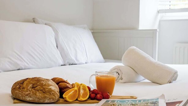 Ein ordentlich bezogenes Bett mit weißen Leinentüchern, ein Brotlaib, Orangenscheiben, Erdbeeren und ein Glas Orangensaft darauf.
