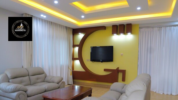 A living room with beige sofas, a wooden coffee table, and a TV mounted on the wall. - Residence Six Sigma (Entebbe)