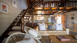 A spacious living area with a high ceiling, a wooden staircase, a beige sofa, wicker chairs, and a coffee table. - Karongwe - Noka House (Hoedspruit)
