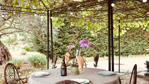Un espacio de comedor al aire libre con una pérgola, mesas dispuestas con platos, copas de vino y una botella.