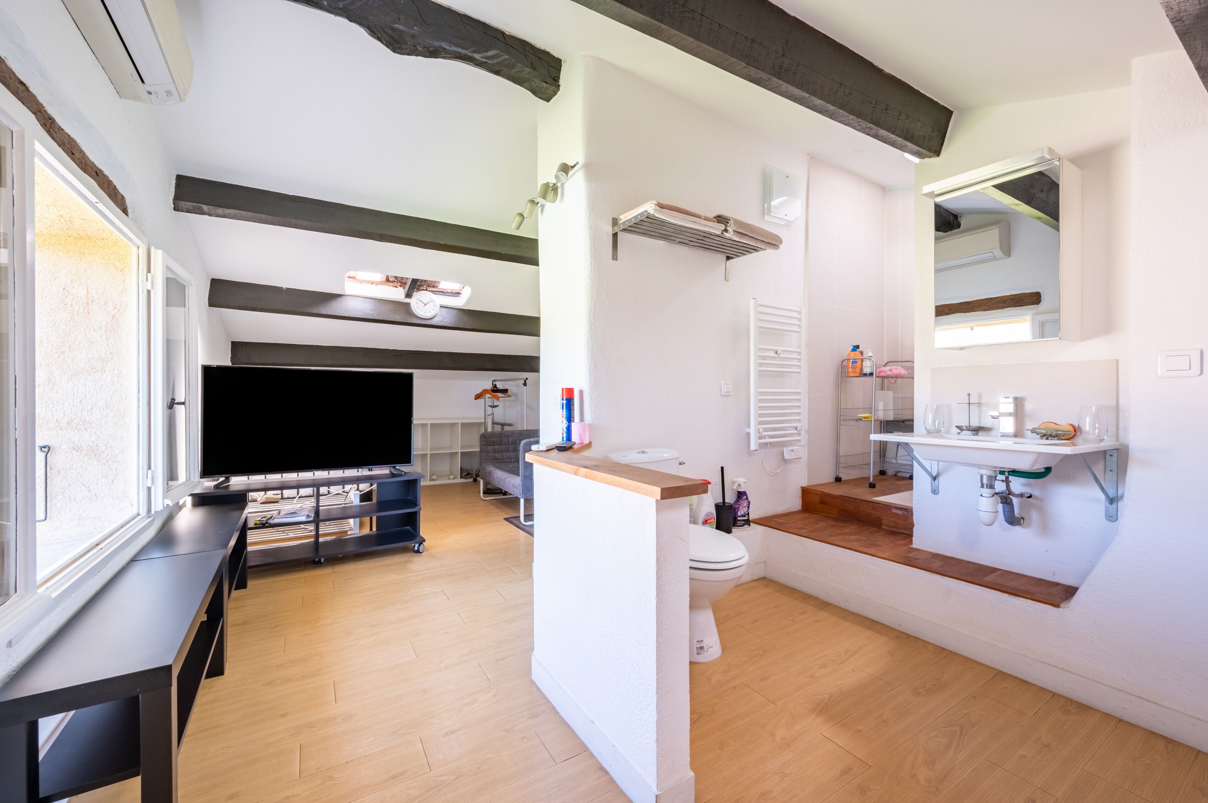 Ein modernes Badezimmer mit Toilette, Waschbecken und Spiegel. Ein Fenster sorgt für natürliches Licht, und im angrenzenden Raum befindet sich ein Fernseher.