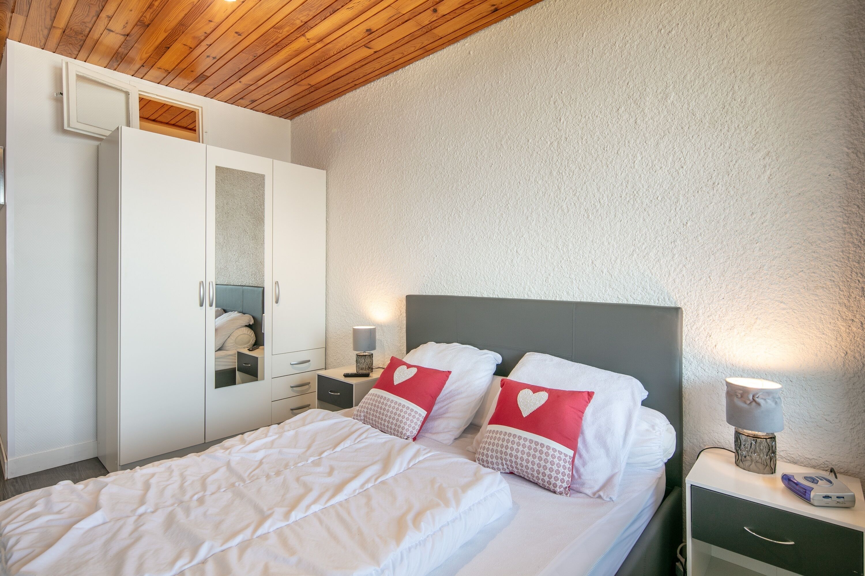 A bedroom with a bed, white wardrobe, bedside tables, and a wooden ceiling.