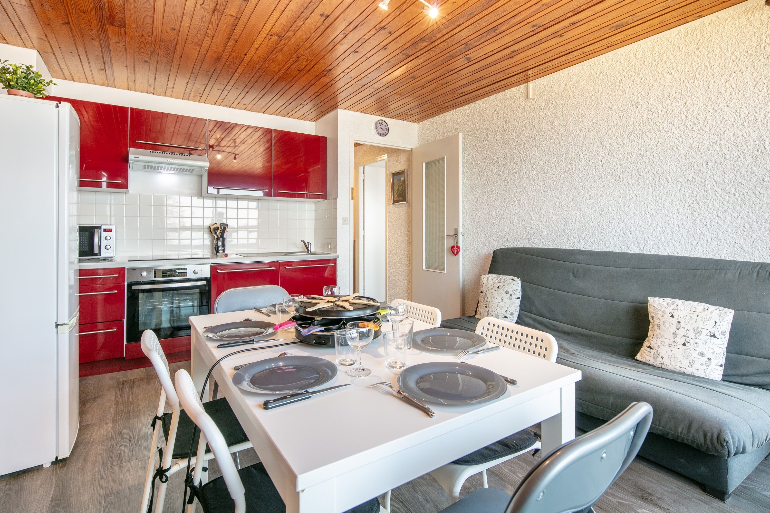 A modern kitchen with red cabinets, a white dining table set for two, and a grey sofa in the living area.