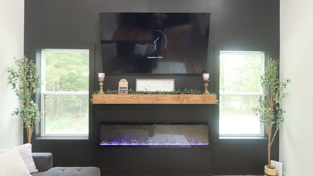 Ein modernes Wohnzimmer mit einem großen Flachbildfernseher an einer schwarzen Wand, einem Kamin darunter und einem Holztisch mit Tablett und Pflanze.