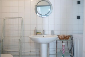 Une salle de bain avec un lavabo blanc, un miroir rond, une cabine de douche en verre et une petite étagère contenant des articles de toilette.