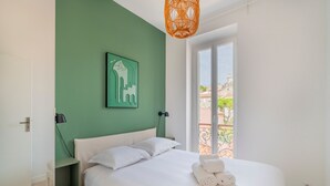 A neatly made bed with white linens, a green accent wall, a framed artwork, and a balcony with a wrought iron railing. - Monte Bello - Charmant Appt 2 personnes (Marseille)