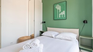 A neatly made bed with white linens and rolled towels, a green wall with a framed artwork, and a bedside table with a lamp. - Monte Bello - Charmant Appt 2 personnes (Marseille)