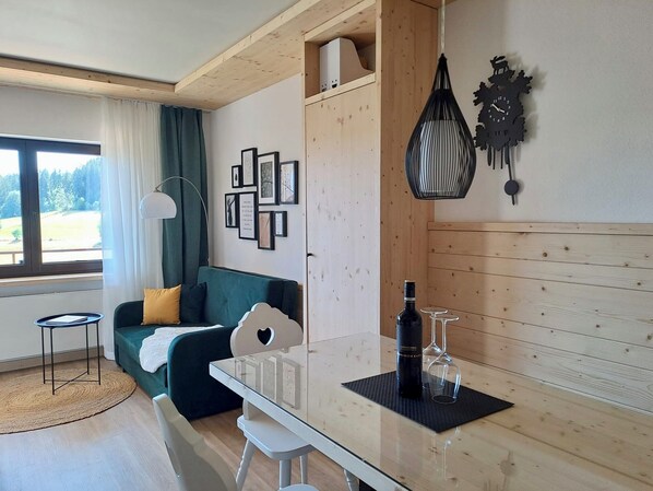 A modern living room with a green sofa, a small white table, and a wooden cabinet. There are framed pictures on the wall and a unique clock. - Vacation apartment "Das Waldo" with a view of the Black Forest (Lenzkirch)