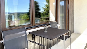 Een balkon met een tafel en stoelen, grote ramen bieden uitzicht op groen en een verafgelegen landschap.