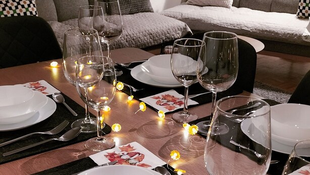 A dining table set with plates, cutlery, and wine glasses, surrounded by candles and a sofa in the background.