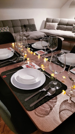 A dining table set with plates, cutlery, and wine glasses, surrounded by candles and a sofa in the background.