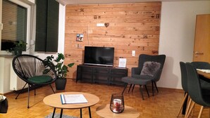 A cozy living room with a wooden wall, a flat-screen TV, a black sofa, a green chair, and a small table with a book.