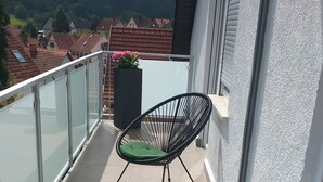 A balcony with two black wire-framed chairs with green cushions, a glass railing, and a view of a hill with houses.