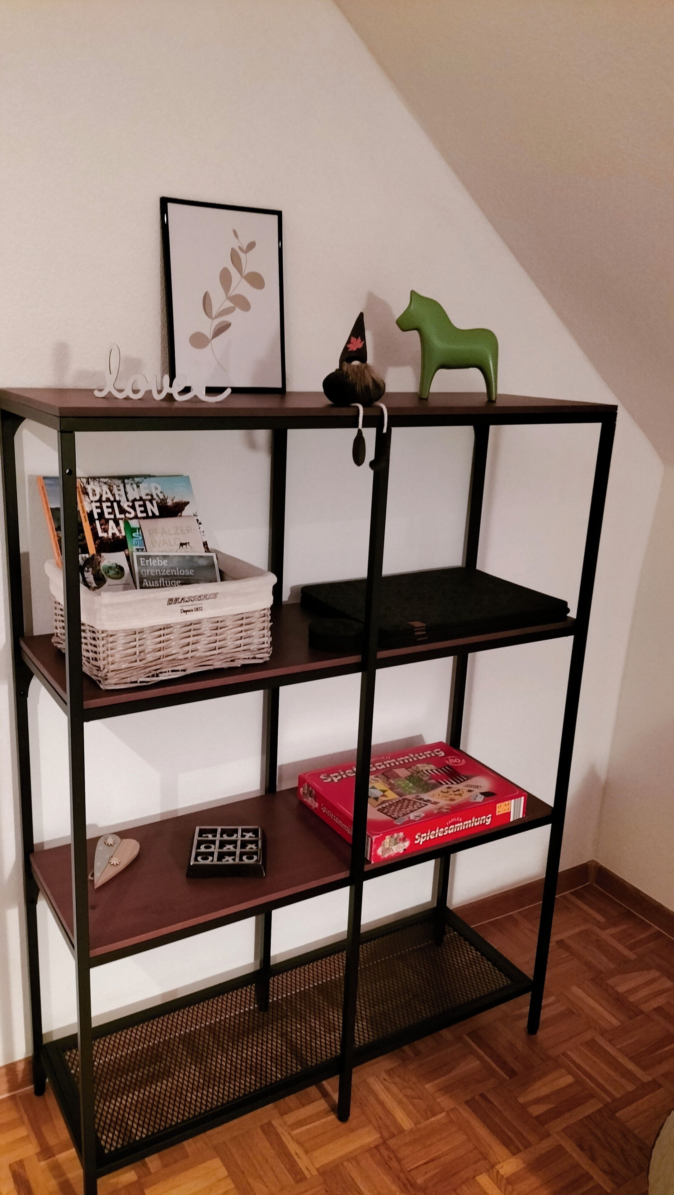 A black metal shelf unit with three shelves, a wicker basket, a red board game, and a framed picture with leaves.