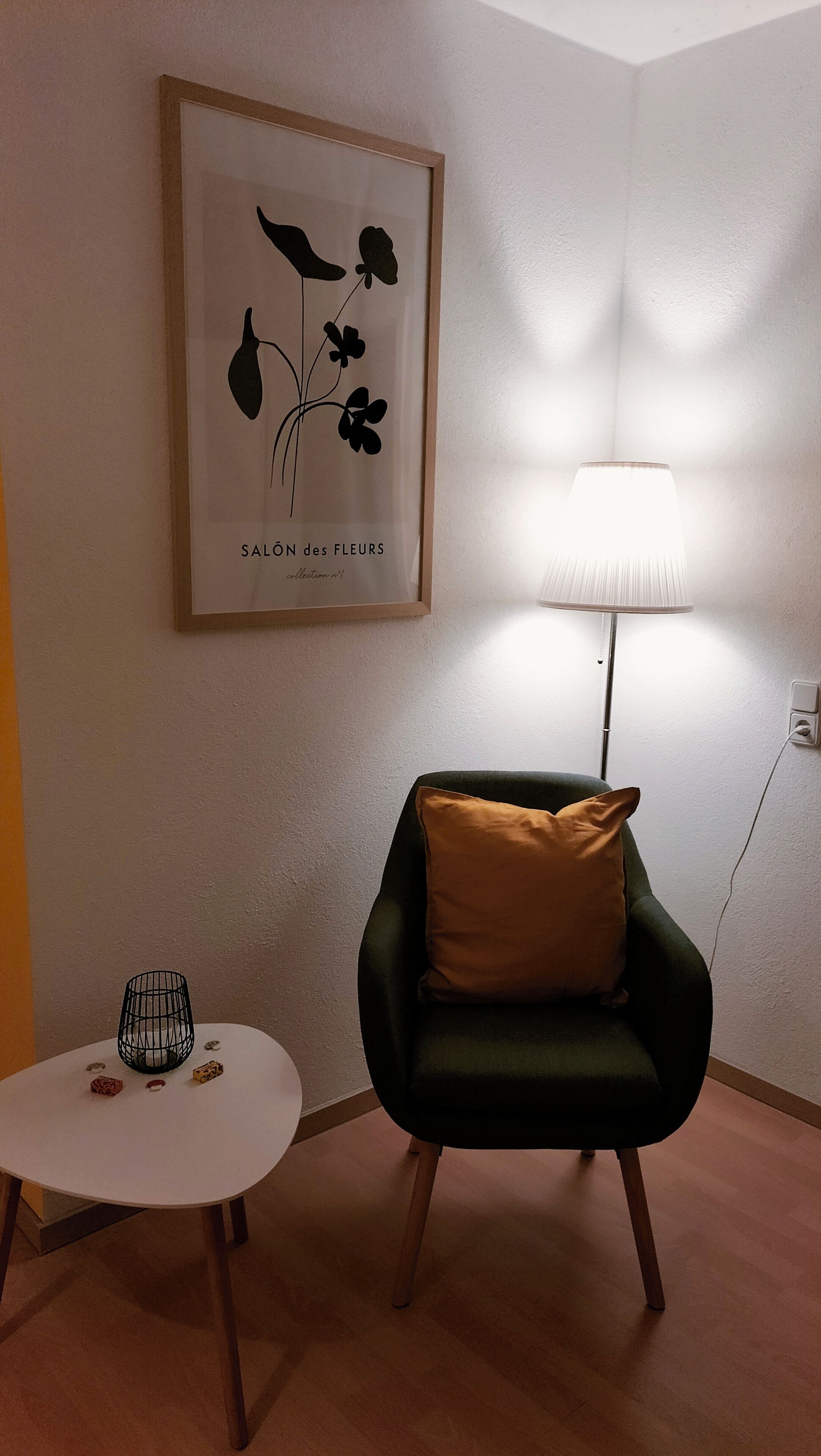 A room with a framed artwork, a lamp, a chair, a small table, and a floor lamp.