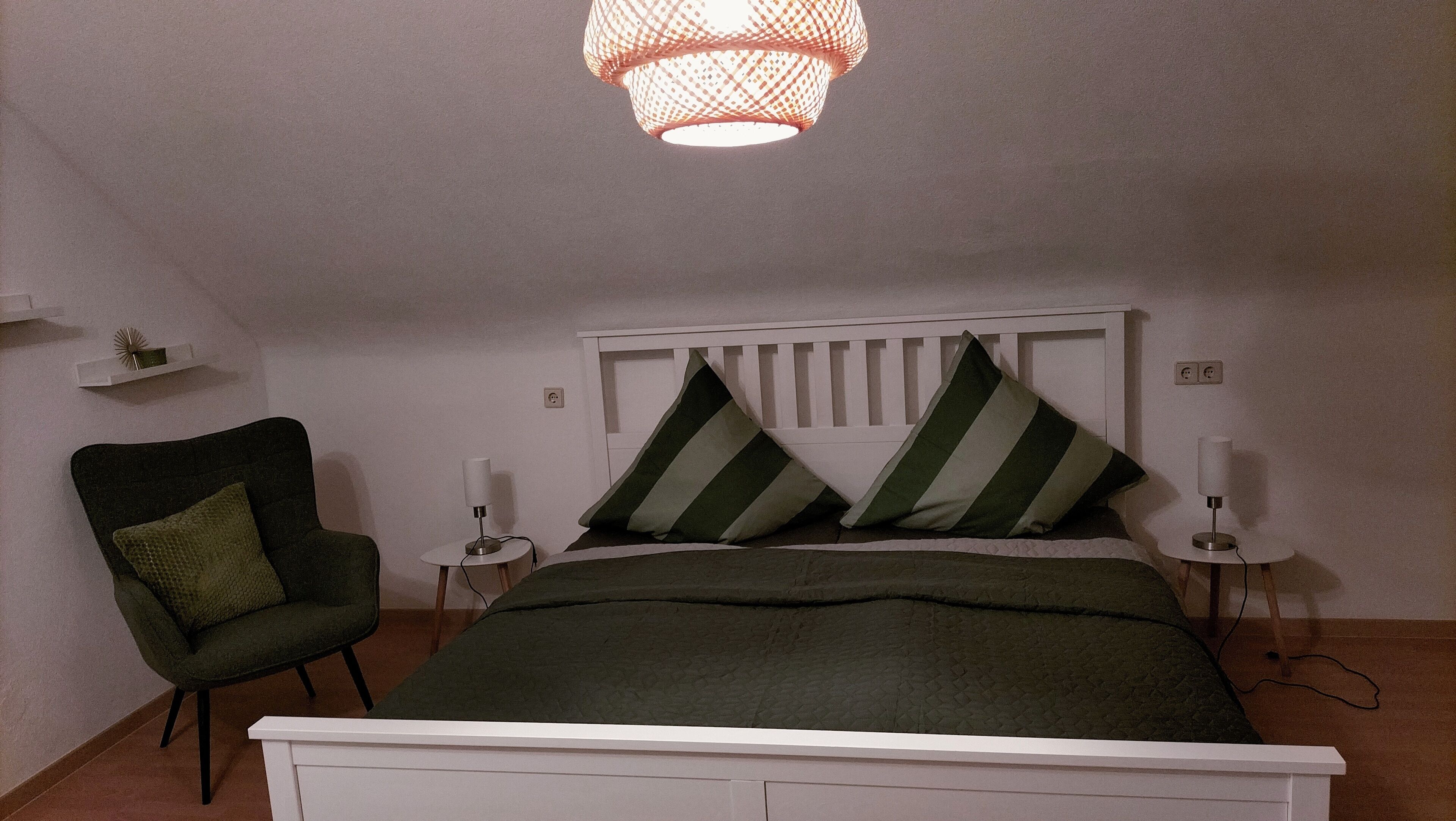 Un dormitorio con una cama blanca, almohadillas verdes a rayas, un sillón verde, lámparas de noche y una lámpara colgante.