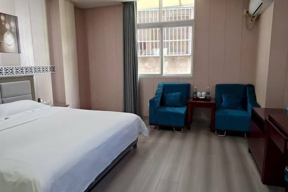 A hotel room with a bed, two blue armchairs, a small table with a bottle, and a window with bars.