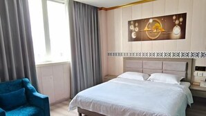 A hotel room with a bed, a blue armchair, a wall-mounted artwork, and a window with curtains.