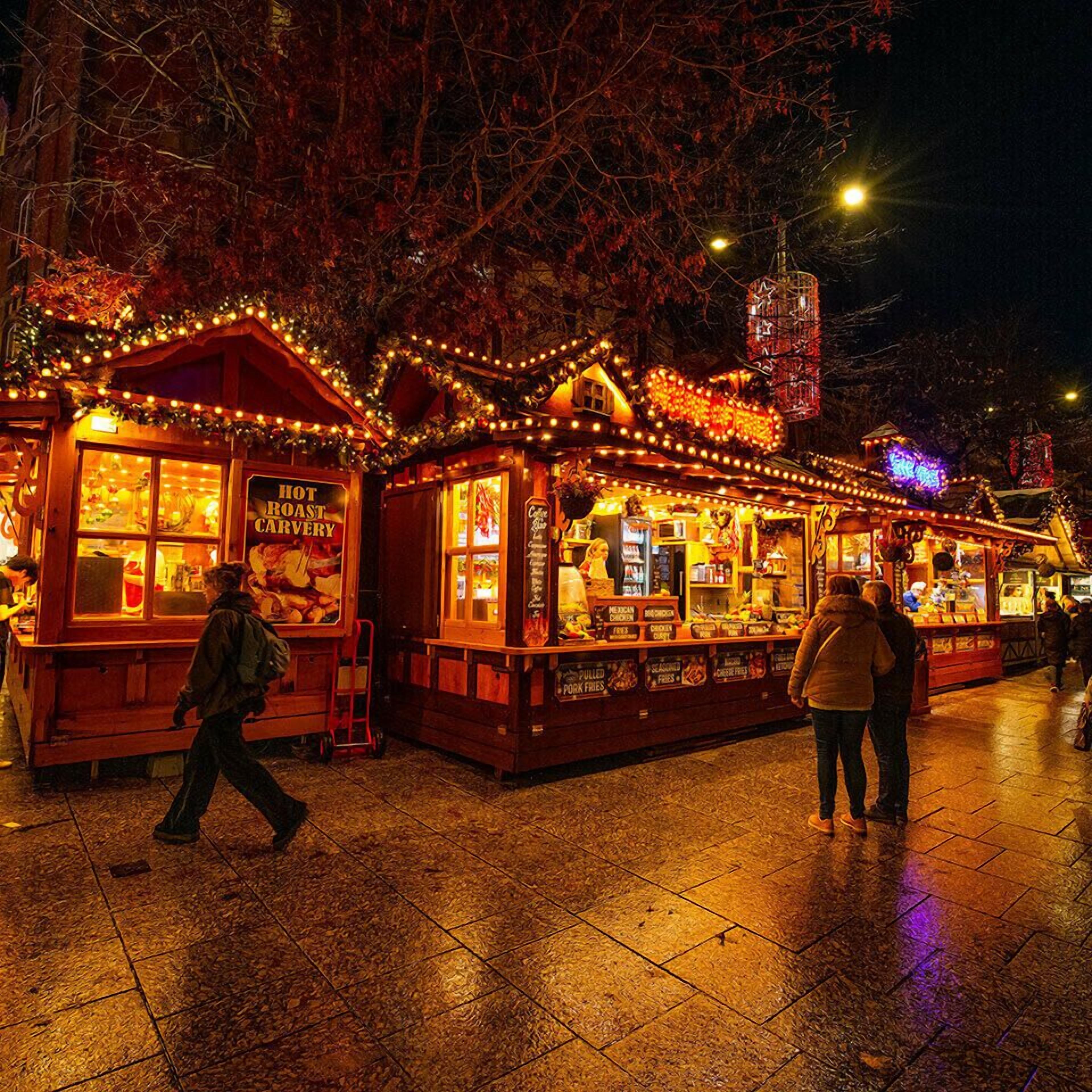 A nighttime market with illuminated stalls, including a hot roast coffee shop and a food vendor selling Mexican chicken and pulled pork fries.