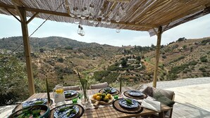 Mesa posta para dois, com pratos, copos e jarra, com vista para uma paisagem montanhosa.