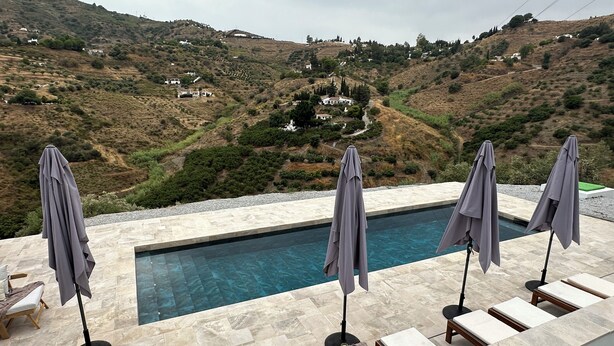 Área da piscina com espreguiçadeiras e guarda-sóis, com vista para uma paisagem montanhosa com casas.