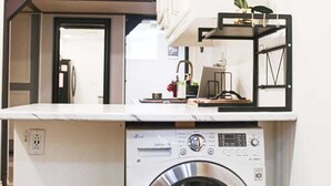 A compact laundry area with a washing machine and a drying rack, a hanging light fixture, and a small shelf with a potted plant. - Ideal for long stays, Near/airport GYM-Parking. (Toronto)